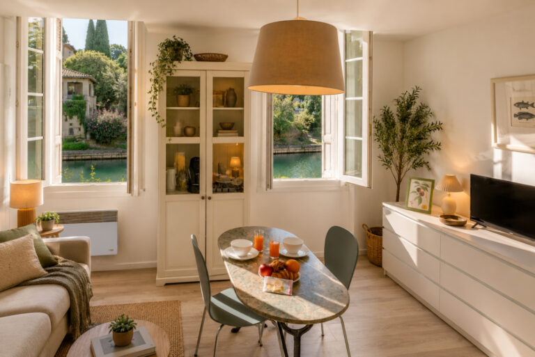 Salon lumineux avec canapé, télé et une table dressée au centre, décoration chaleureuse, vue sur le canal par les fenêtres.