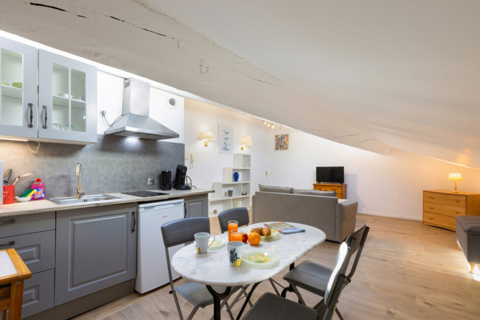 Cuisine équipée ouverte sur un vaste espace de vie sous combles avec une table dressée en petit déjeuner