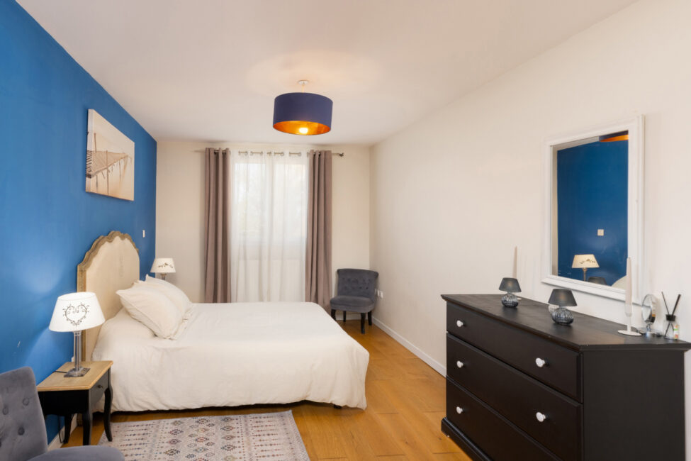 Photographe immobilier à Carcassonne : quand un shooting professionnel change tout 9 Chambre avec lit double, mur bleu, commode et lumière naturelle.