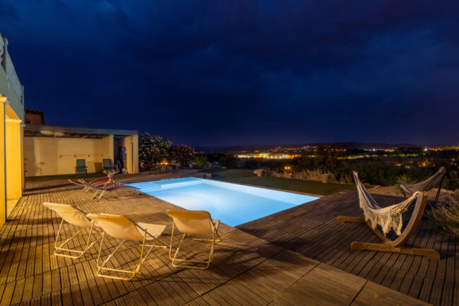 Piscine et terrasse en bois éclairées avec vue sur les lumières de la ville.