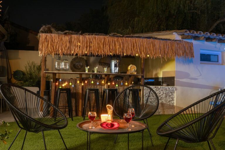 Espace détente extérieur de nuit avec bar couvert et éclairé, fauteuils noirs, table basse, carafe et verres disposés pour créer une ambiance conviviale.