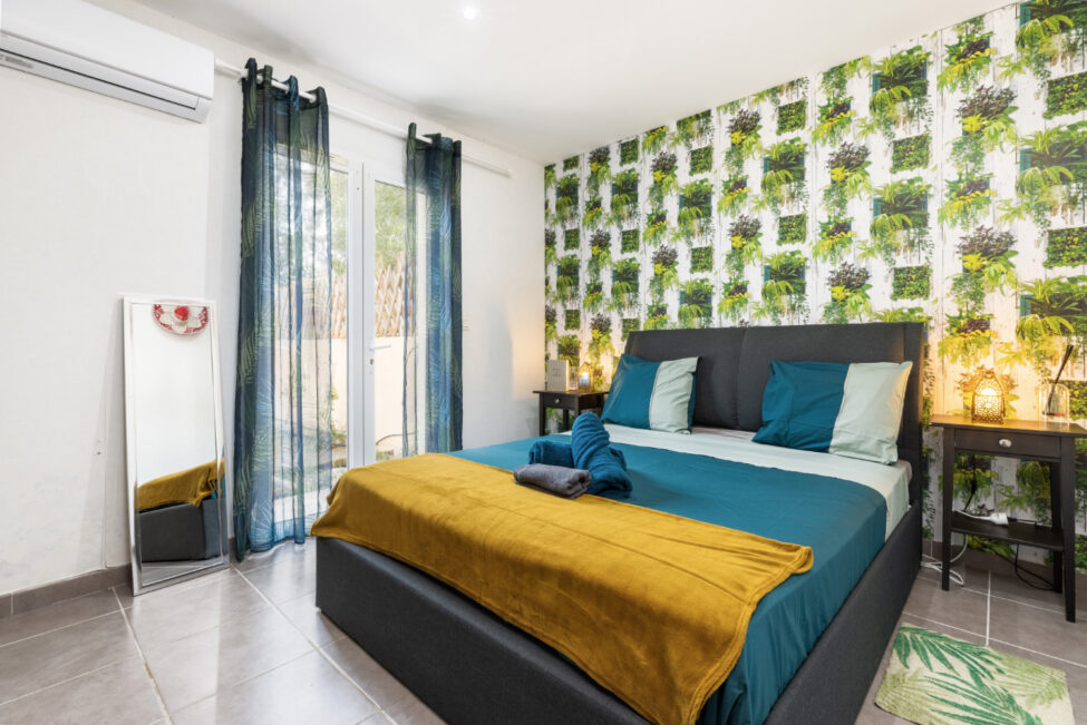 Chambre double lumineuse avec linge de lit bleu et jaune, mur à motif végétal, tables de chevet, miroir et accès direct vers l’extérieur.