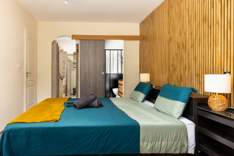 Chambre double avec linge de lit bleu et vert, mur décoratif en tasseaux de bois, lampes de chevet et accès ouvert vers le dressing et la salle de bain.