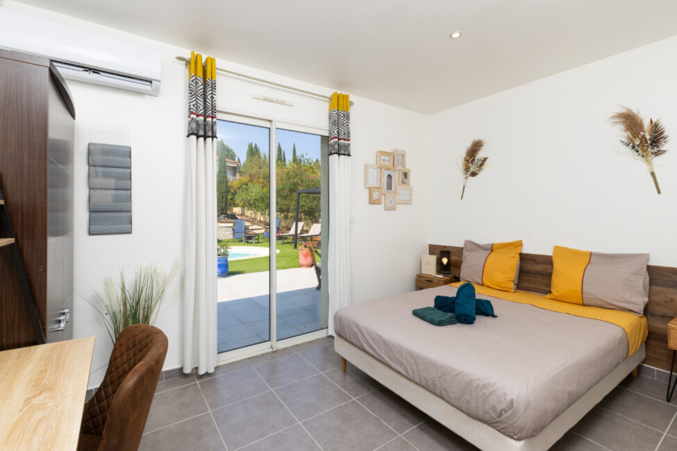Chambre double lumineuse avec linge de lit beige et jaune, bureau, décoration murale et baie vitrée donnant directement sur le jardin et la piscine.