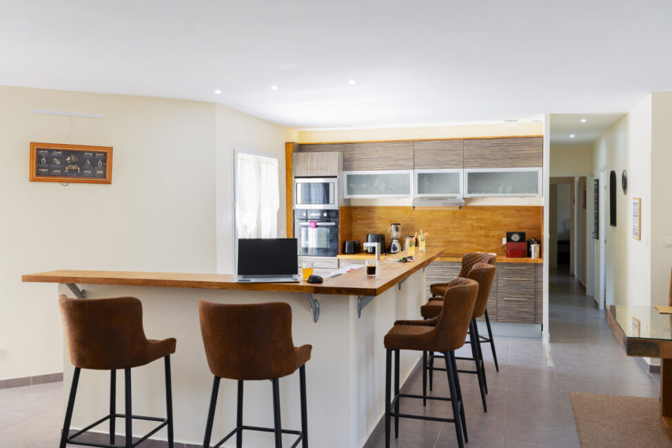 Cuisine ouverte équipée avec grand comptoir en bois, plusieurs tabourets hauts, électroménager intégré et rangements.