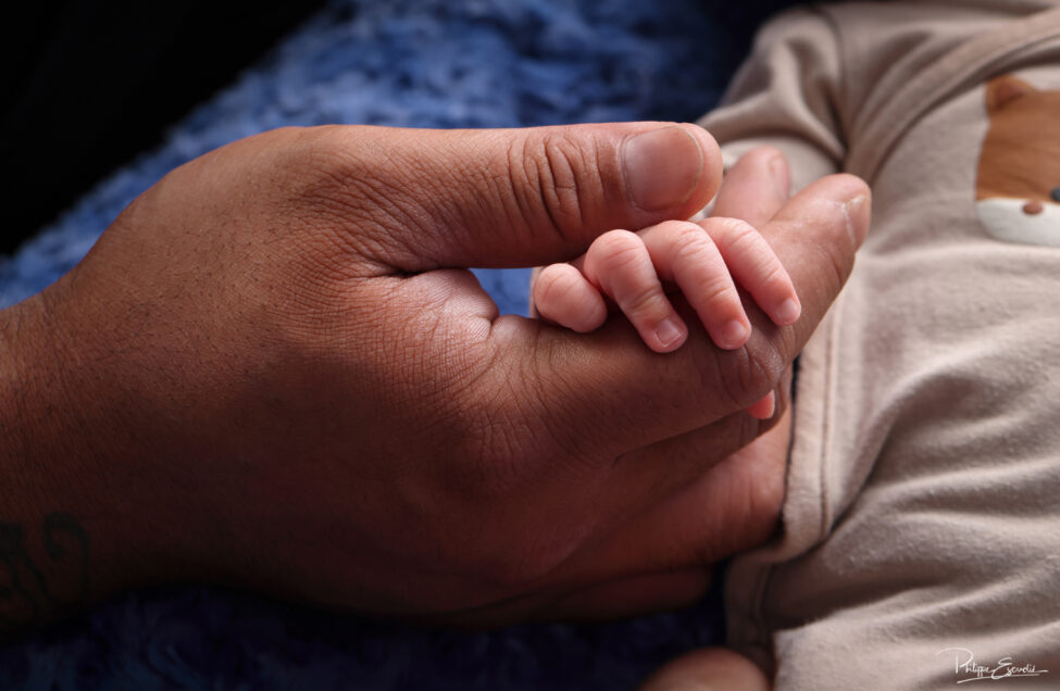 Shooting photo d’un bébé du bout du monde 7 Gros plan sur la petite main d’un nouveau-né posée sur la main de son papa, mettant en valeur le contraste entre leurs mains.