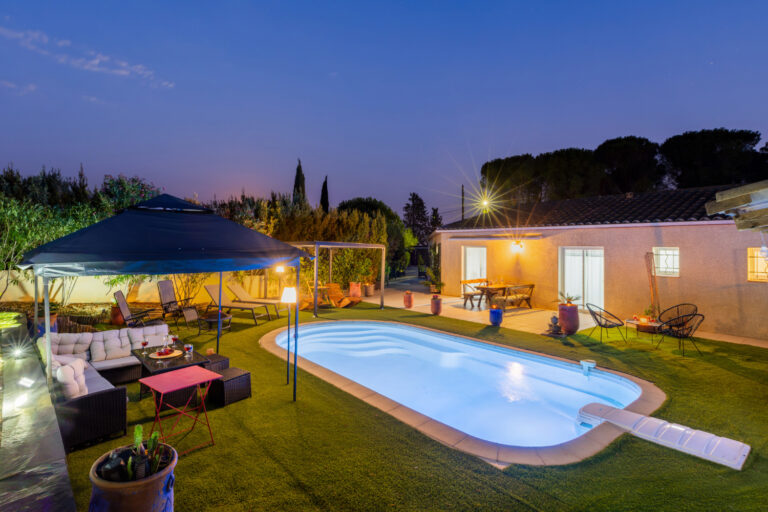 Piscine illuminée au crépuscule devant la maison et sa terrasse, avec salon de jardin, transats et différents espaces de détente aménagés.