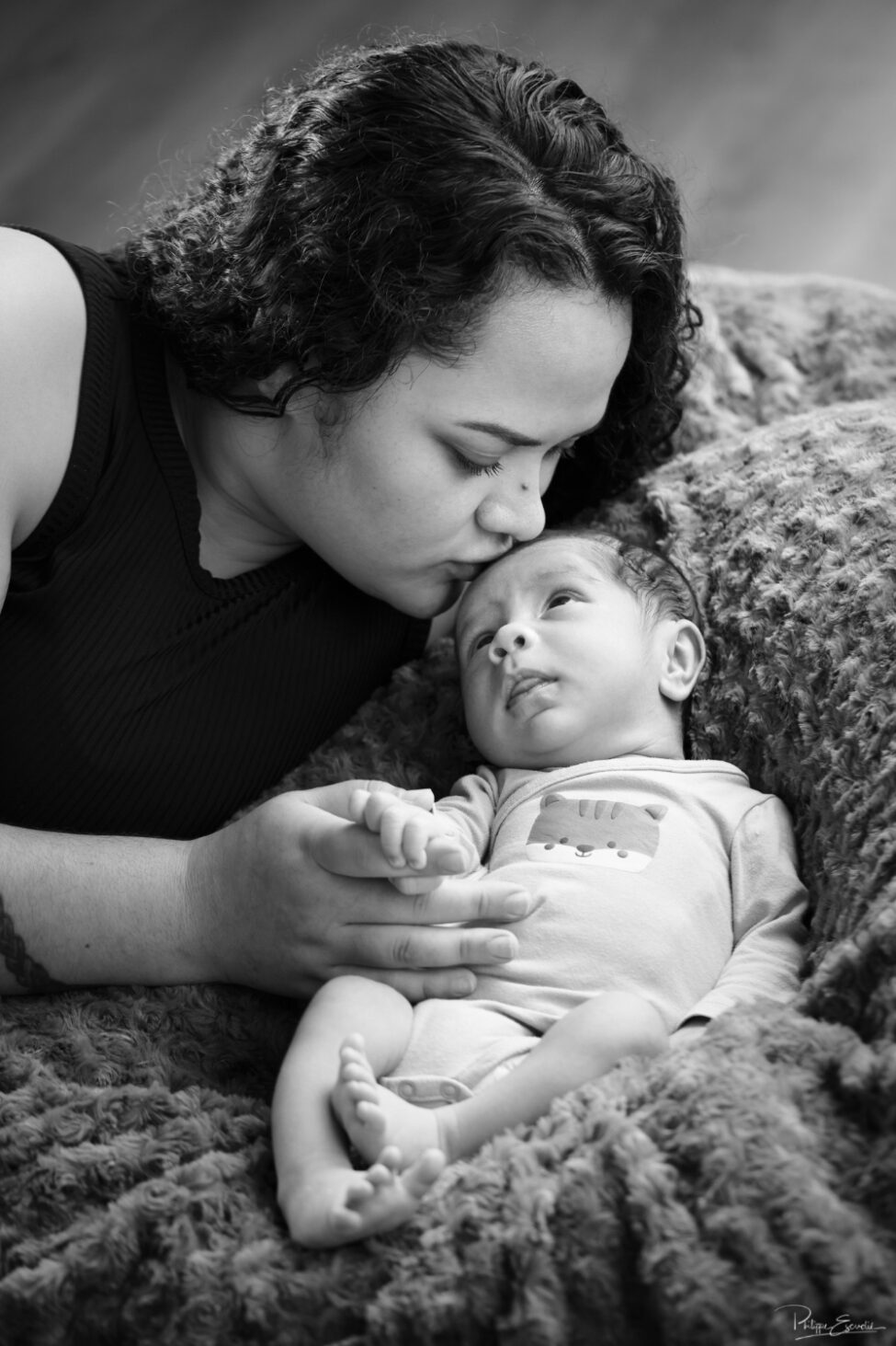 Shooting photo d’un bébé du bout du monde 6 Portrait en noir et blanc d’une maman embrassant tendrement son nouveau-né allongé contre elle sur une couverture douce.