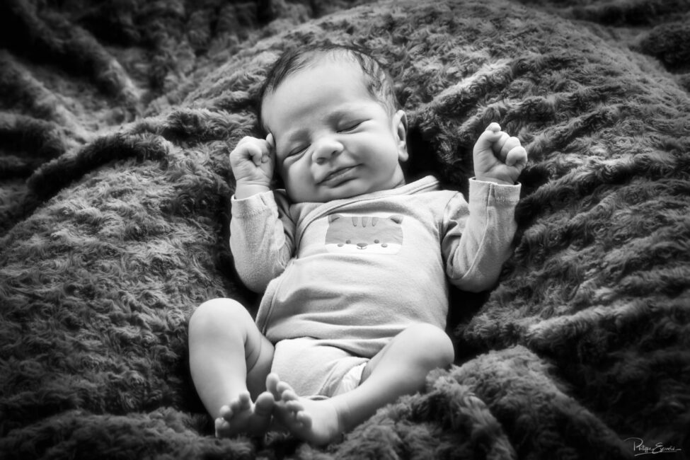 Shooting photo d’un bébé du bout du monde 2 Nouveau-né photographié en noir et blanc, allongé sur une couverture douce, les yeux fermés avec une expression souriante.