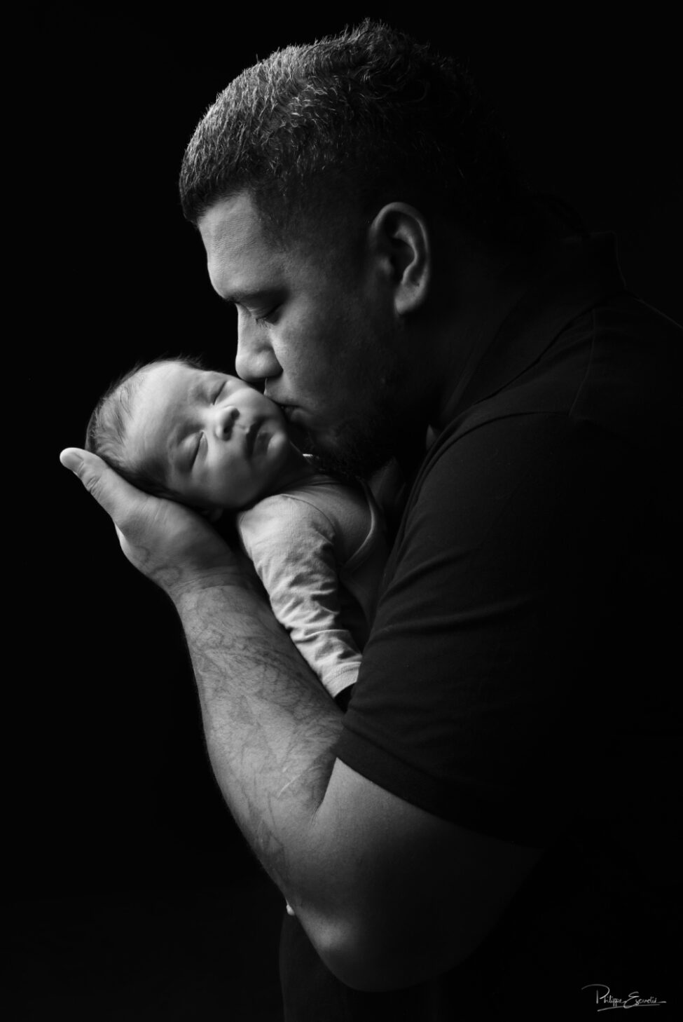 Shooting photo d’un bébé du bout du monde 5 Portrait en noir et blanc d’un papa tenant son nouveau-né endormi dans ses bras et l’embrassant doucement sur la joue.