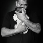Portrait en noir et blanc d’un papa tenant son nouveau-né contre lui et l’embrassant sur la tête, sur un fond sombre.