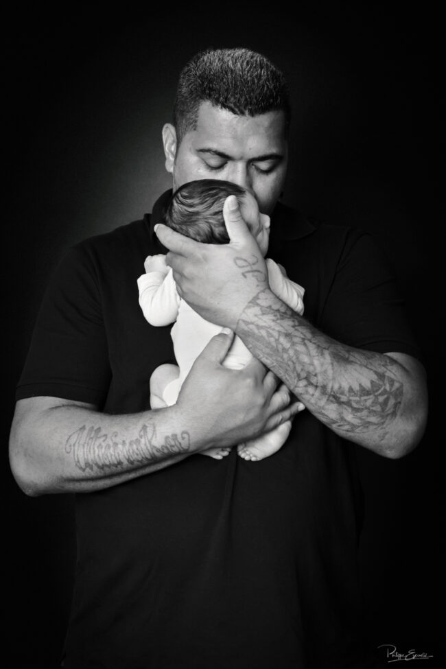 Portrait en noir et blanc d’un papa tenant son nouveau-né contre lui et l’embrassant sur la tête, sur un fond sombre.