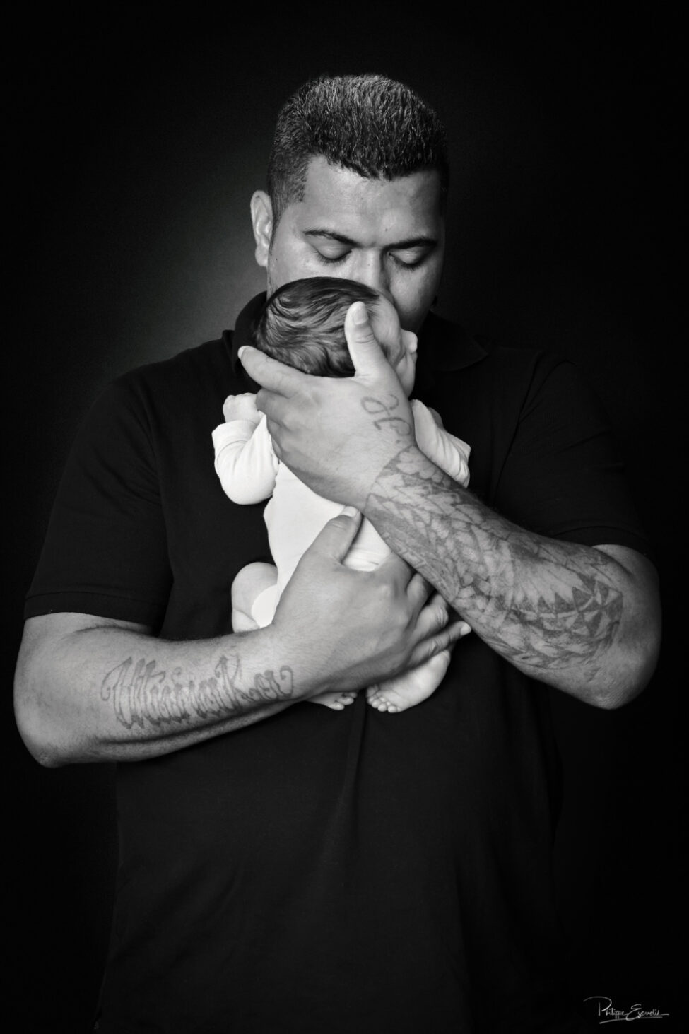 Shooting photo d’un bébé du bout du monde 4 Portrait en noir et blanc d’un papa tenant son nouveau-né contre lui et l’embrassant sur la tête, sur un fond sombre.