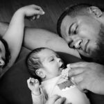 Portrait familial en noir et blanc montrant un nouveau-né allongé entre ses parents qui le regardent et lui tiennent délicatement les mains.