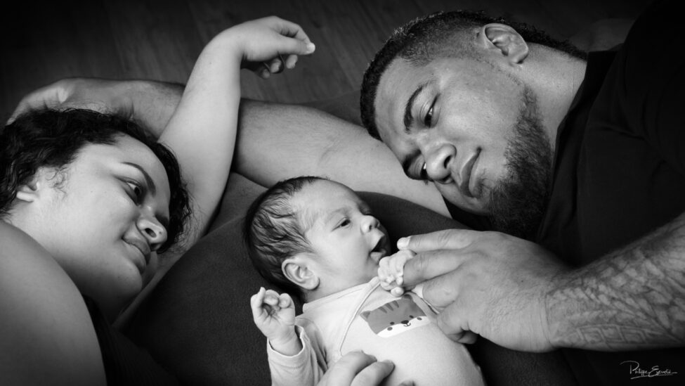 Shooting photo d’un bébé du bout du monde 3 Portrait familial en noir et blanc montrant un nouveau-né allongé entre ses parents qui le regardent et lui tiennent délicatement les mains.
