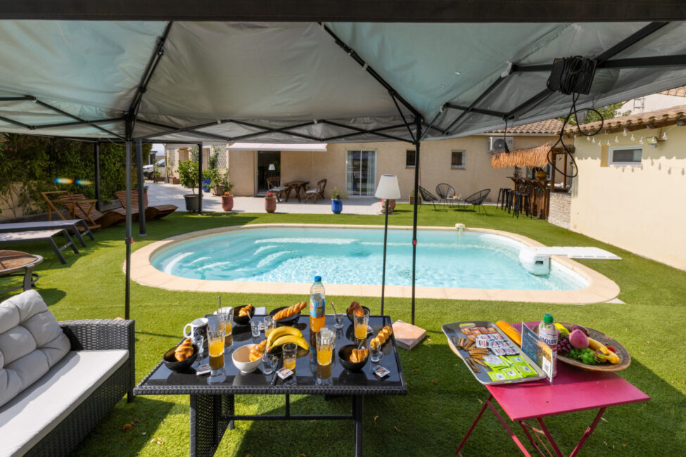 Petit-déjeuner dressé sous une tonnelle face à la piscine, avec boissons, viennoiseries et fruits sur les tables.