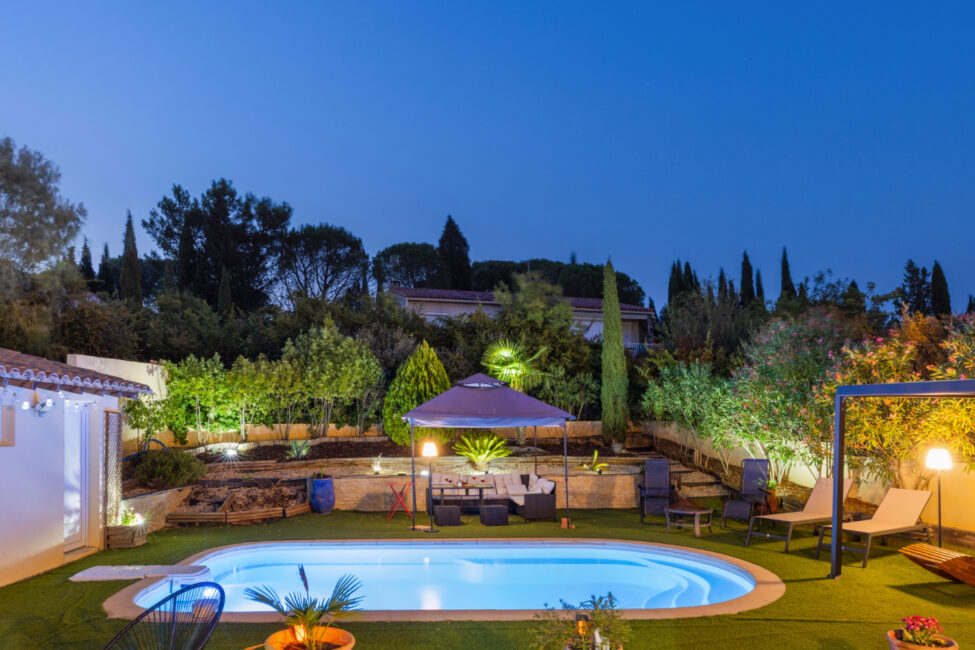 Vue de nuit sur une piscine éclairée entourée d’un jardin paysager, avec salon extérieur sous tonnelle, transats et éclairages d’ambiance.