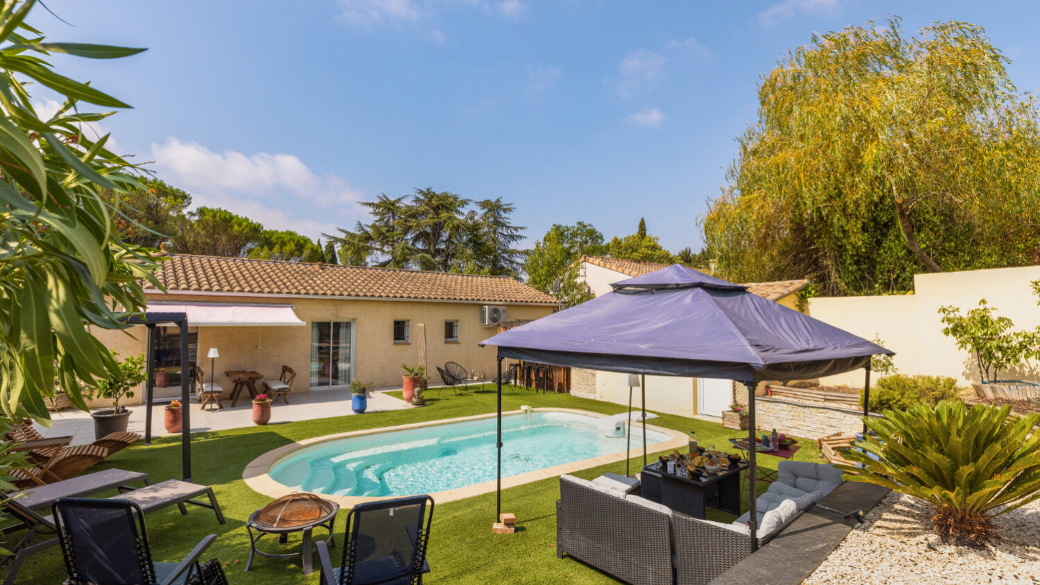 Vue d’ensemble de la piscine et du jardin avec salon extérieur sous tonnelle, transats et maison en arrière-plan.
