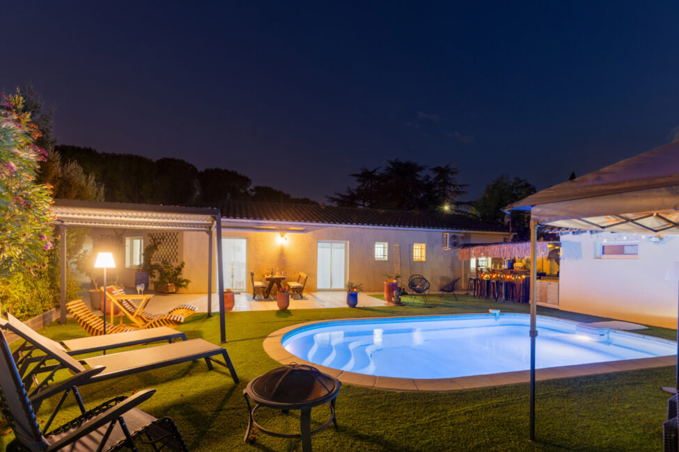 Piscine éclairée de nuit face à la maison, avec terrasse aménagée, transats, pergola, espace bar et éclairages extérieurs.