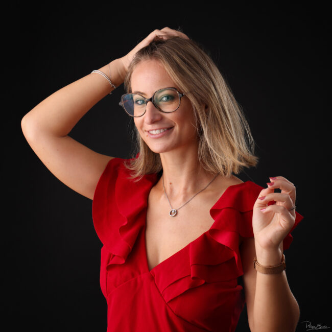 Portrait rapproché d’une femme en tenue rouge, une main de les cheveux, lors d’un shooting en studio sur fond sombre.