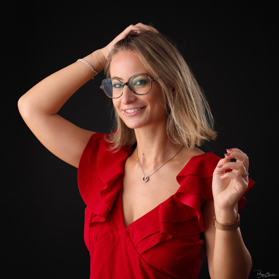 Portrait rapproché d’une femme en tenue rouge, une main de les cheveux, lors d’un shooting en studio sur fond sombre.
