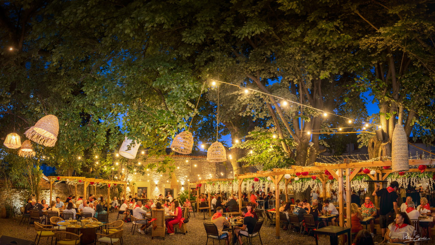 reportage-photo-soiree-ambiance-restaurant Vue d’ensemble d’une soirée festive dans un restaurant en extérieur. les gens sont sous des pergolas en bois avec des lampions.