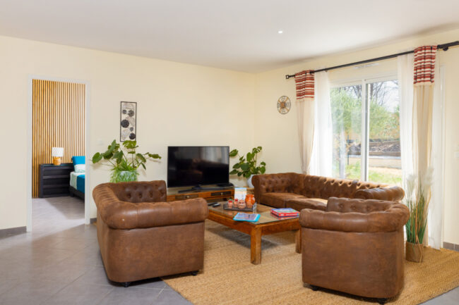 Salon lumineux aménagé avec deux canapés et deux fauteuils en cuir, table basse, télévision, plantes et grande baie vitrée.