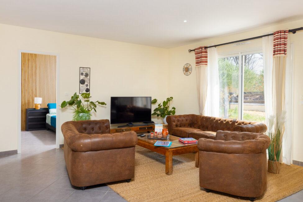 Salon lumineux aménagé avec deux canapés et deux fauteuils en cuir, table basse, télévision, plantes et grande baie vitrée.