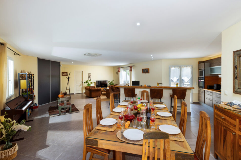 Grande pièce de vie ouverte avec table à manger dressée, cuisine avec comptoir et tabourets, salon et piano.