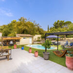 Vue d’ensemble de la terrasse et du jardin avec piscine, pergola, transats en bois, espace bar, mobilier extérieur et végétation arborée.