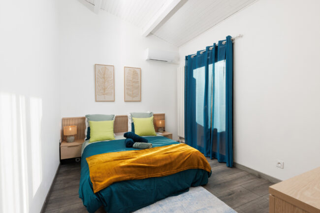 Une chambre accueillante avec lit queen, mobilier en bois clair et linge de lit bleu et ocre, dans un décor lumineux aux lignes épurées.