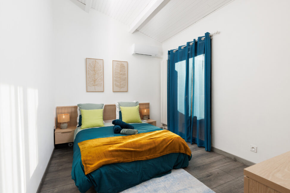 Une chambre accueillante avec lit queen, mobilier en bois clair et linge de lit bleu et ocre, dans un décor lumineux aux lignes épurées.