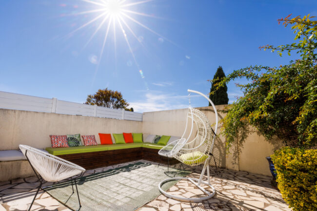 Un espace de détente extérieur avec banquettes colorées, fauteuil suspendu et végétation, dans une cour aménagée sous le soleil.