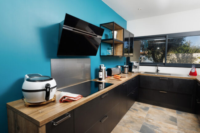 Une cuisine équipée aux meubles noirs et plans de travail en bois, mise en valeur par un mur bleu et une large fenêtre donnant sur le jardin.