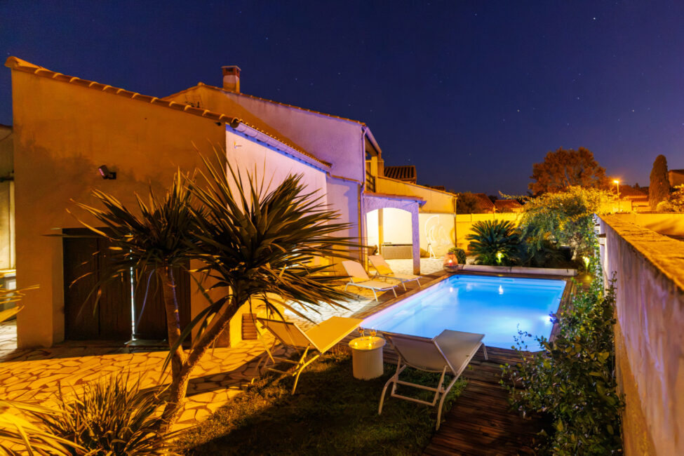 Une vue nocturne de la maison et de sa piscine, où les éclairages chaleureux de la façade contrastent avec le bleu lumineux de l'eau.