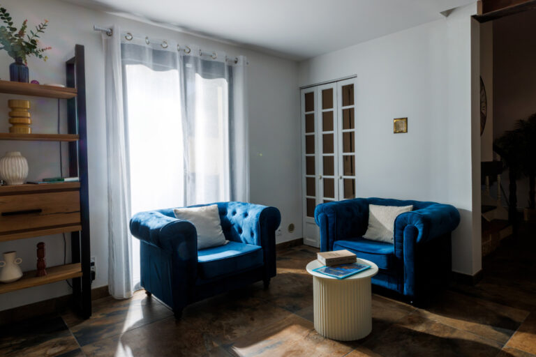 Deux fauteuils en velours bleu accompagnés d'une table basse blanche composent un coin salon confortable, éclairé par la lumière naturelle.