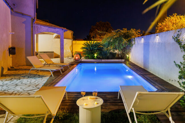 Deux transats installés face à une piscine illuminée, accompagnés d'une petite table et de verres, dans une ambiance nocturne intime.