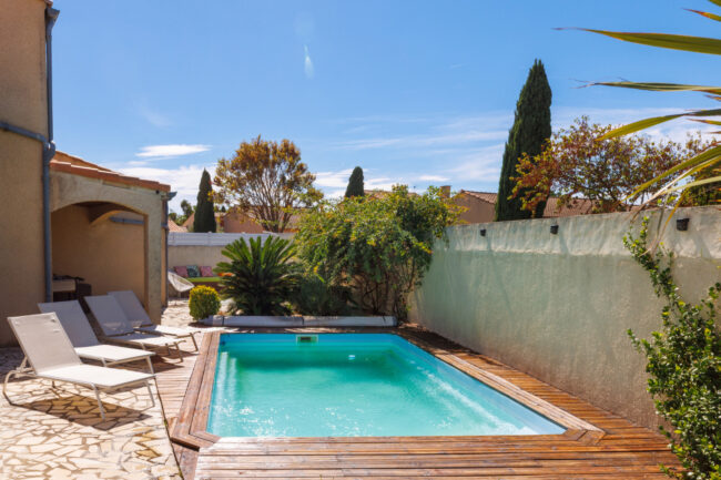 Une piscine bordée d'une terrasse en bois, entourée de végétation et de transats, dans un espace extérieur ensoleillé.