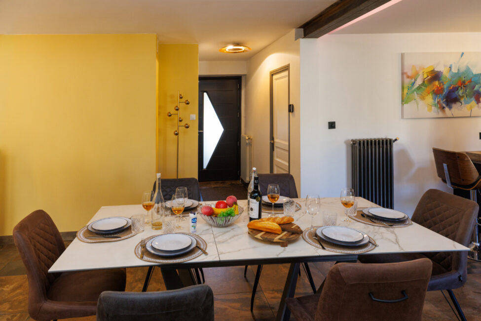 Une grande table dressée pour six personnes, entourée de fauteuils confortables, dans une salle à manger aux couleurs chaleureuses.
