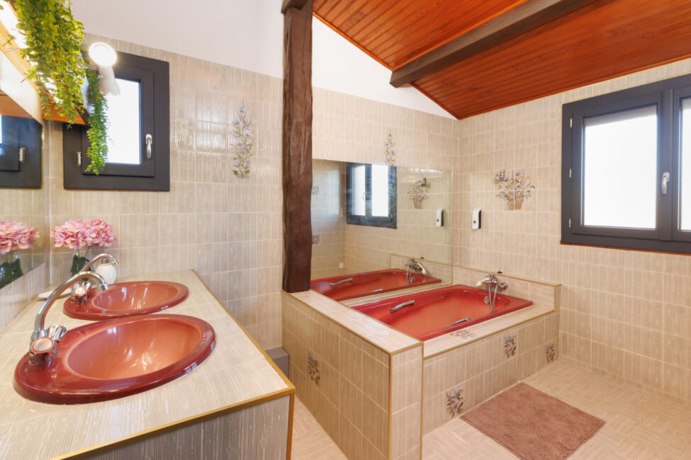 Une salle de bains avec baignoire encastrée, deux vasques aux tons cuivrés et carrelage à motifs floraux, sous un plafond habillé de bois.