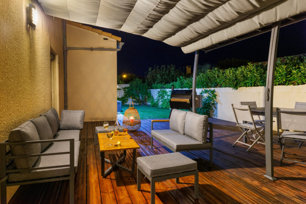 Un salon extérieur aménagé sous une terrasse couverte, avec mobilier confortable, éclairage chaleureux et vue sur le jardin à la nuit tombée.