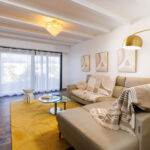 Un salon lumineux avec canapé d'angle beige, table basse en verre et tapis jaune, prolongé par une grande baie vitrée ouverte sur l'extérieur.
