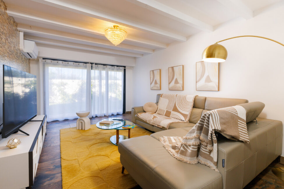 Un salon lumineux avec canapé d'angle beige, table basse en verre et tapis jaune, prolongé par une grande baie vitrée ouverte sur l'extérieur.