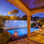 Depuis la terrasse couverte, la piscine illuminée se dévoile dans une atmosphère de début de soirée, entourée de transats et de végétation.
