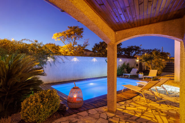 Depuis la terrasse couverte, la piscine illuminée se dévoile dans une atmosphère de début de soirée, entourée de transats et de végétation.