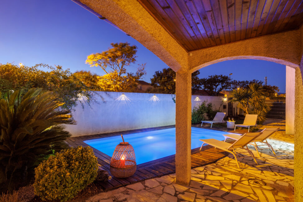 Depuis la terrasse couverte, la piscine illuminée se dévoile dans une atmosphère de début de soirée, entourée de transats et de végétation.
